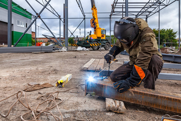 Man welder in welding mask, building uniform and blue protective gloves brews metal welding machine on street construction, in the background construction site - Photo, Image