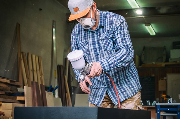Carpintero experimentado en ropa de trabajo y pequeño propietario de buiness pinta una caja de madera de la cómoda en color negro en el taller, en el fondo una gran cantidad de herramientas
 - Foto, imagen