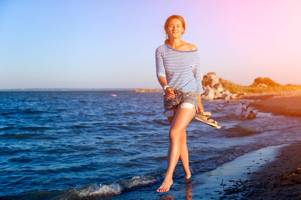 Una donna dai capelli scuri allegra sorride, cammina lungo la spiaggia, calcia le onde, spruzza molto e gode del sole luminoso in una giornata estiva. Concetto di vacanze estive al mare e stile dal vivo
 - Foto, immagini