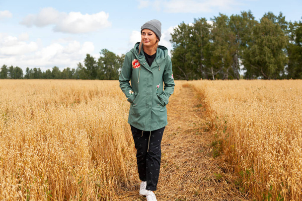 Pretty fresh darkhaired young woman walking outdoors in the sun and high grass  field and wearing a coat, knitting hat, jeans.Concept of autumn  holidays at village  and live style - Fotó, kép