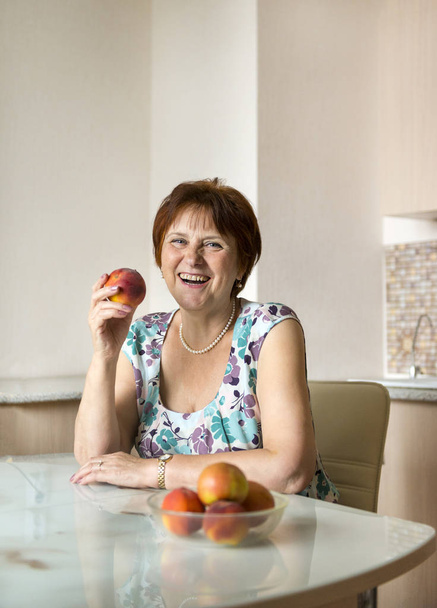 adult woman with fruit in the kitchen, woman laughing, woman eating fruit, apples, peaches - Foto, Imagen