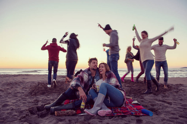 Junges Paar genießt mit Freunden am Lagerfeuer am Strand bei Sonnenuntergang Bier trinken - Foto, Bild