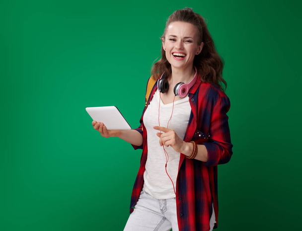 smiling modern student woman in red shirt with backpack and headphones holding tablet PC and pointing aside while posing on green background - Foto, Imagen