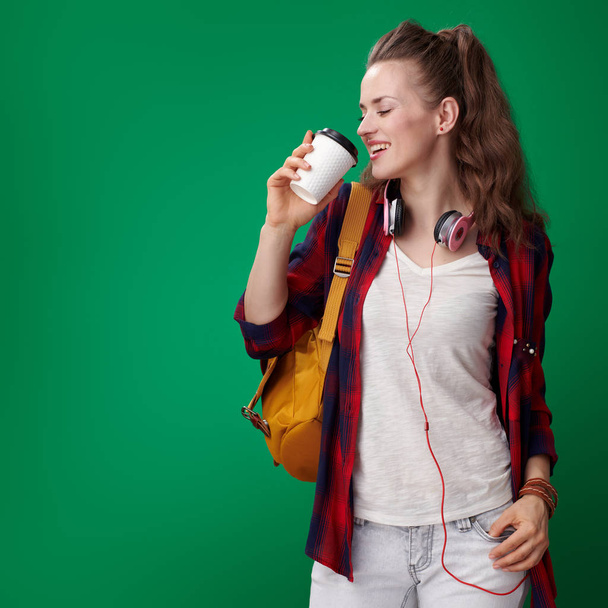щаслива молода студентка в червоній сорочці з рюкзаком та навушниками, що п'ють каву на зеленому фоні
 - Фото, зображення