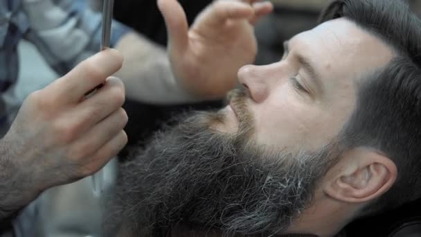 Barber shaves men with a long beard with straight razor blade in s hair salon or barbershop. Mans haircut and shaving at the hairdresser, barber shop and shaving salon. - Πλάνα, βίντεο