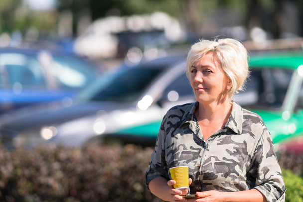 Smiling happy lady wearing camouflage shirt while holding small orange cup with multiple blurred cars in the background - Photo, Image