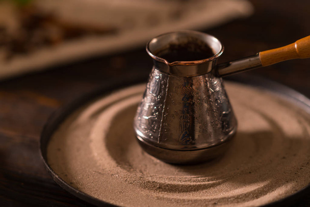 Engraved metal coffee pot with a wooden handle standing on a ceramic stand in shadowy light in the kitchen - Foto, Imagem