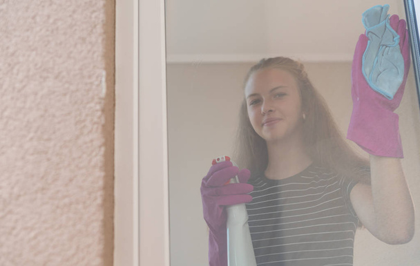 junge Frau lächelt, während sie ein Fenster mit einem Spray und Tuch reinigt, das durch die Glasscheibe gesehen wird - Foto, Bild