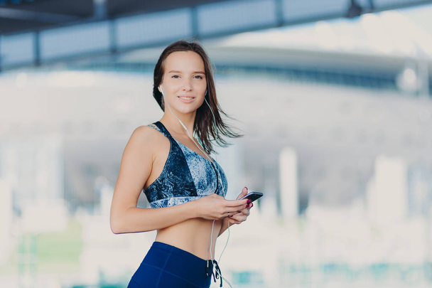 Active Caucasian woman with dark hair, healthy skin, dressed in sportswear, uses up to date gadget, listens music with earphones, has charming smile, stands outdoor. People and lifestyle concept - Foto, afbeelding
