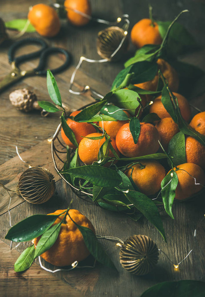 Fresh ripe tangerines with leaves in tray, decoration toys, scissors and light garland over rustic wooden table background - Photo, image
