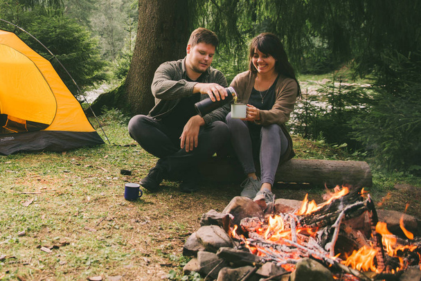 coppia seduta vicino al fuoco e bere tè caldo. concetto di trekking. auto con tenda sullo sfondo
 - Foto, immagini