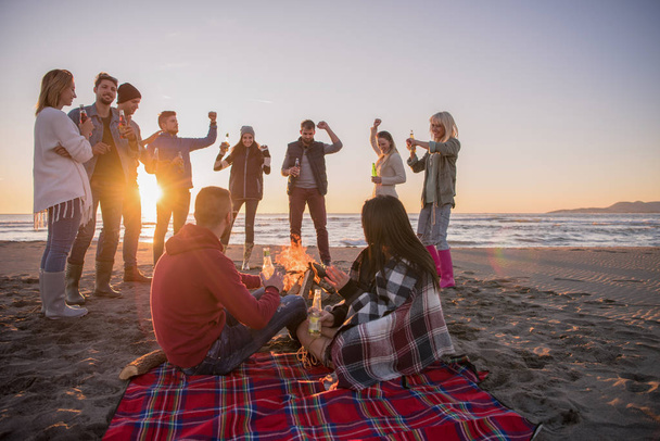 ビーチでキャンプファイヤーの周りの友人と楽しむ若いカップル日没時にビールを飲む - 写真・画像