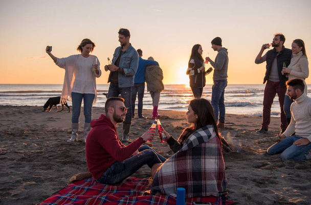 Młoda para zabawia się z przyjaciółmi przy ognisku na plaży o zachodzie słońca pijąc piwo - Zdjęcie, obraz
