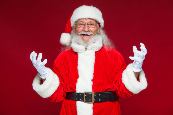 Christmas. Good Santa Claus in white gloves shows faces, grimaces, shows his tongue. Not standard behavior. Isolated on red background. - Photo, image