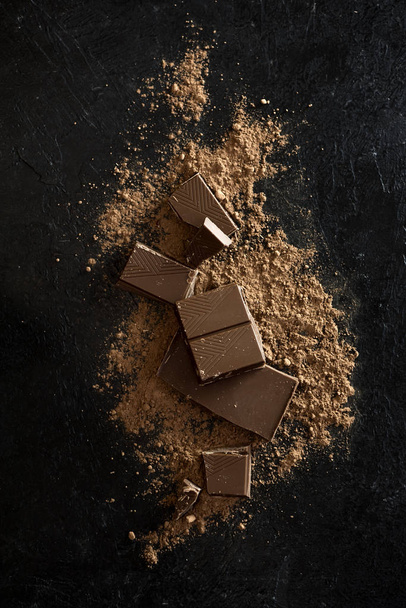 Broken chocolate pieces and cocoa powder on black. Chunks of chocolate onr dark stone background. - Photo, image