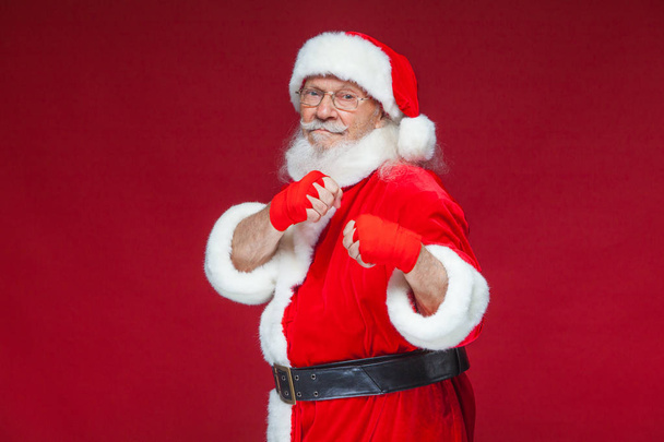Christmas. Santa Claus with red bandages wound on his hands for boxing imitates kicks. Kickboxing, karate, boxing. Isolated on red background. - Zdjęcie, obraz