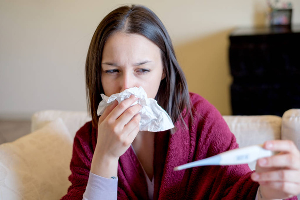 Mujer enferma cubierta con una manta en el sofá con fiebre alta y una gripe comprobando la temperatura corporal
 - Foto, Imagen