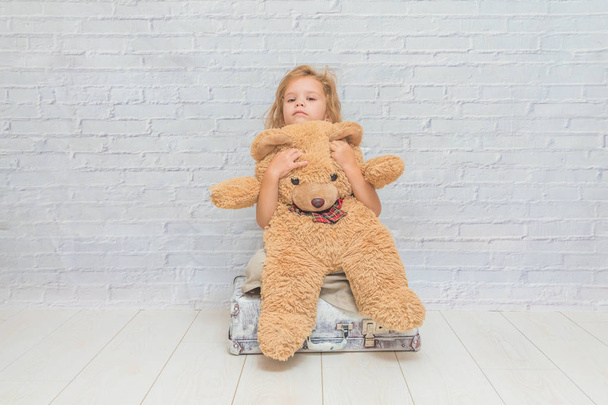 girl, baby in dress on white brick wall background with suitcase, toy bear - Foto, immagini