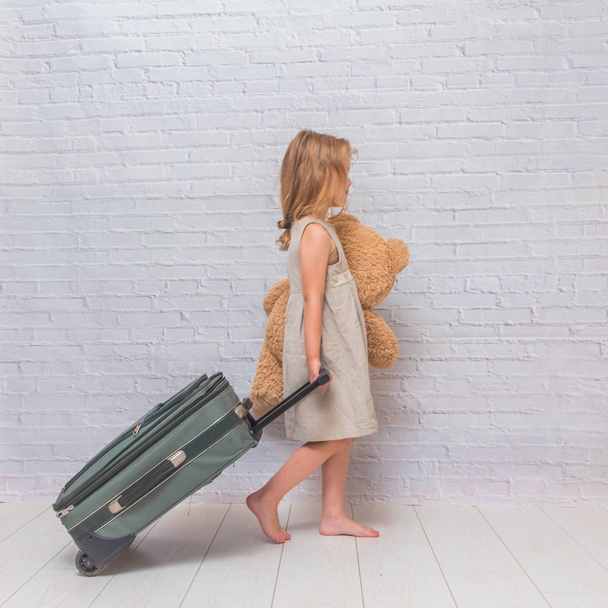 girl, baby in dress on white brick wall background with suitcase, toy bear - Foto, Imagem