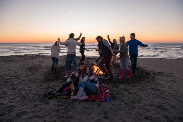 Junges Paar genießt mit Freunden am Lagerfeuer am Strand bei Sonnenuntergang Bier trinken - Foto, Bild