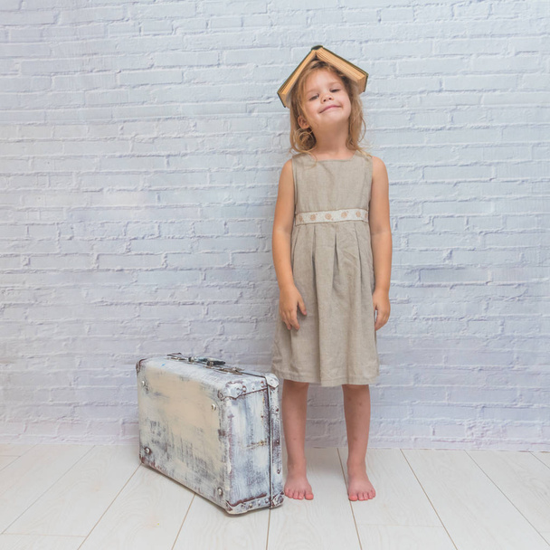 girl, baby in dress on white brick wall background with suitcase, with a book - Zdjęcie, obraz