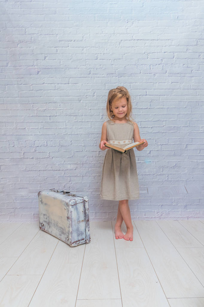 girl, baby in dress on white brick wall background with suitcase, with a book - Foto, imagen