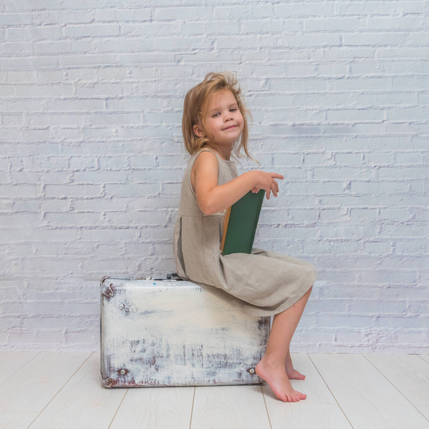 Kız, bebek elbise bavul, bir kitapla beyaz tuğla duvar zemin üzerine - Fotoğraf, Görsel