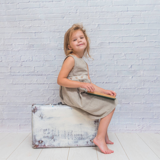 niña, bebé en vestido sobre fondo de pared de ladrillo blanco con maleta, con un libro
 - Foto, imagen