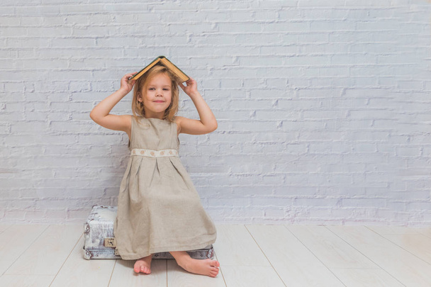 girl, baby in dress on white brick wall background with suitcase, with a book - Fotoğraf, Görsel