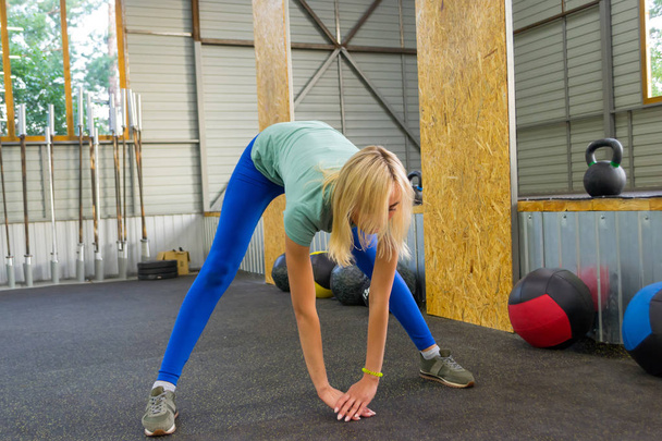 The blond girl in blue leggings in the gym is training, leaning forward downwards, touching the floor with the arms and head down on the background of sports equipment: medical balls,weights and bars. - Photo, Image
