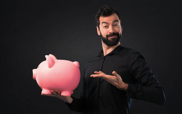 Jóképű férfi szakállal gazdaság egy fekete háttér piggybank - Fotó, kép