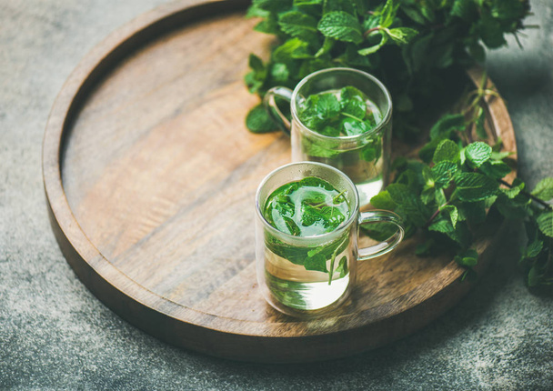 Hot herbal mint tea drink in glass mugs with fresh garden mint leaves over wooden tray over concrete background - Fotoğraf, Görsel