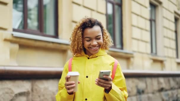 Attractive mixed race woman is using modern smartphone looking at screen and smiling surfing the net or watching photos and holding take away coffee standing outdoors. - Záběry, video