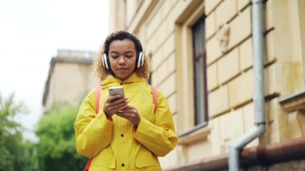 Dolly shot of cheerful young lady listening to music with headphones, singing and using smartphone walking outdoors in modern city. Fun, people and technology concept. - Metraje, vídeo
