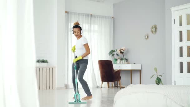 Happy housewife is doing housework and listening to music wearing modern wireless headphones, young lady is singing in mop and dancing having fun at home. - Záběry, video
