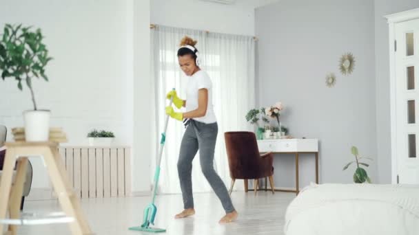 Happy African American woman is washing floor with plastic mop, singing and dancing listening to music through headphones. People and modern technology concept. - Video