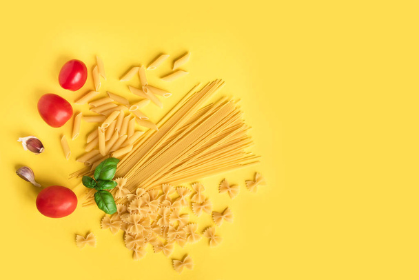 Various uncooked raw pasta with basil,tomatoes and garlic on yellow background. Italian pasta: penne, farfalle, spaghetti. - Foto, Imagem