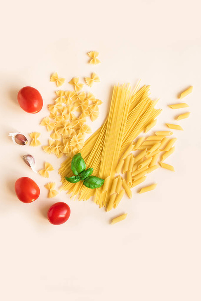 Various uncooked raw pasta with basil,tomatoes and garlic on pink pastel background. Italian pasta: penne, farfalle, spaghetti. - Valokuva, kuva