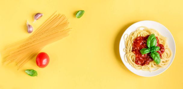 Spaghetti pasta bolognese and ingredients on yellow background, top view. Plate of pasta dish with tomato sauce and basil, creative yellow background. - Photo, Image