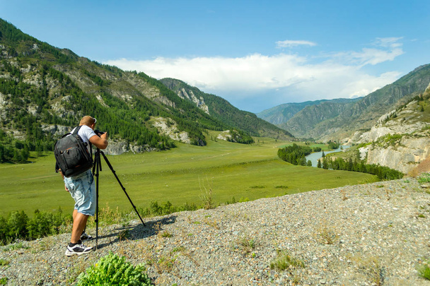 A man photographer is setting up a camera standing on a tripod on top of a rock in the mountains of the Altai makes a photo of a turquoise river of a reel and mountains with clear blue sky and clouds - Photo, Image