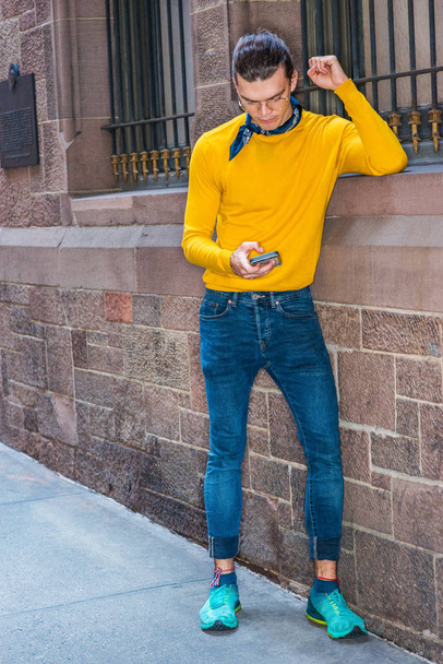 Joven hispano-americano con gafas, camiseta amarilla de manga larga, jeans, zapatillas con estampado verde, bufanda negra pequeña alrededor del cuello, de pie junto a la pared de estilo antiguo en Nueva York, mensajes de texto en el teléfono celular
 - Foto, Imagen