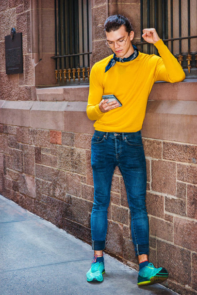 Młodzi Hiszpanie American człowiek nosi okulary, koszula z długim rękawem żółty T, dżinsy, trampki wzorzyste zielony, mały czarny szalik wokół szyi, stojąc przez stary styl ściany w Nowym Jorku, wiadomości tekstowe na telefon komórkowy - Zdjęcie, obraz