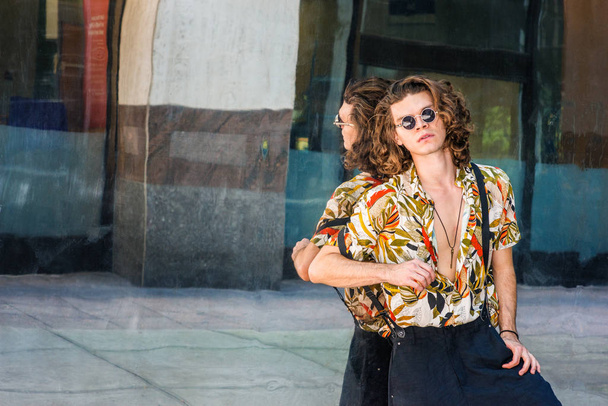 Young Man with brown curly hair, wearing round lenses sunglasses, colorful patterned short sleeve shirt, hanging old key as necklace, pants suspender on shoulder, leaning against mirror in New Yor - Zdjęcie, obraz
