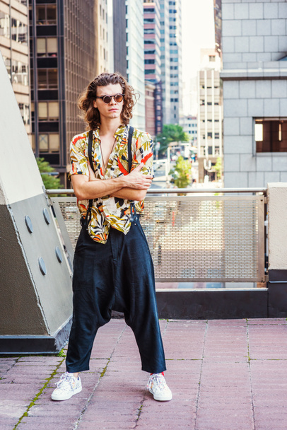 Młody człowiek z brązowe, kręcone włosy, noszenie okularów przeciwsłonecznych, wzorzyste Krótki rękaw koszuli, workowate i luźne spodnie z szelkami, wzorzyste trampki, wiszące stary klucz jako naszyjnik, stojąc na balkonie w Nowym Jorku - Zdjęcie, obraz