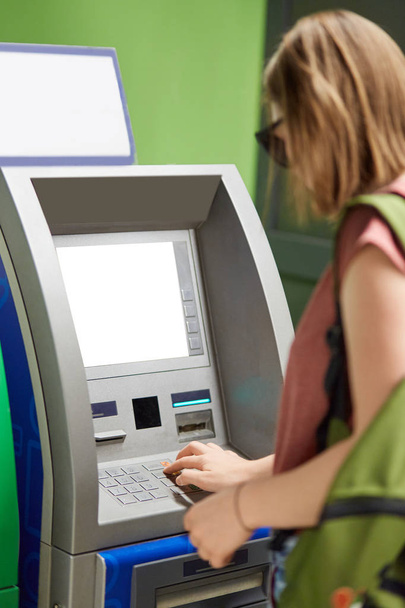 Vertical shot of young fashionable girl stands back near cash machine, withdraws money, types on buttons after inserting credit card, enters pin code, uses ATM machine. Finances and money concept. - Foto, imagen