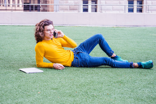 Estudiante joven hispanoamericano universitario con el pelo rizado marrón, con gafas, camiseta amarilla de manga larga, jeans, zapatillas de deporte, sentado hacia atrás en el césped verde en el campus de Nueva York, hablando por teléfono celular
 - Foto, imagen