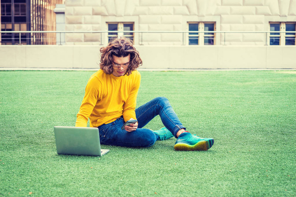 Студент латиноамериканского колледжа учится в Нью-Йорке, с каштановыми кудрявыми волосами, в очках, футболке с желтым длинным рукавом, джинсах, кроссовках, сидит на зеленой лужайке, работает на ноутбуке, пишет смс по телефону
 - Фото, изображение