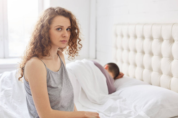 Upset curly female in pyjamas sits on edge of bed, has problems with boyfriend, sort out relationships in bedroom, going to divorce, feels unhappy. Family problems concept. Couple has troubles. - 写真・画像