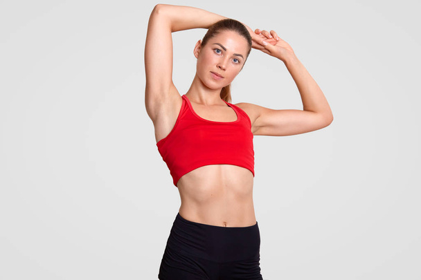 Indoor shot of good looking motivated young sportswoman with perfect body shape, stretches hands, dressed in casual red top, stands against white studio background. People and exercising concept - Fotografie, Obrázek