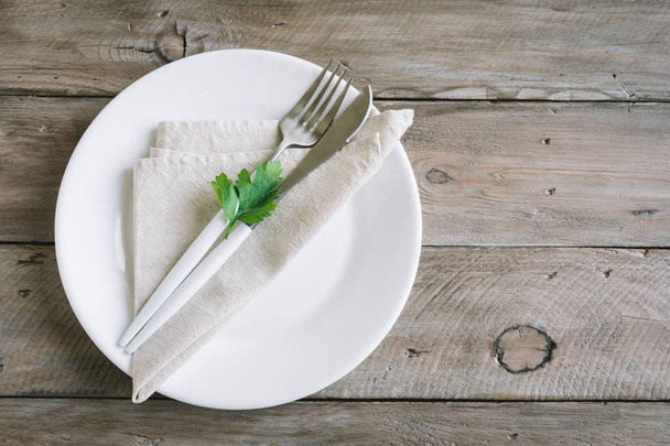 Table setting with white plate, modern cutlery (fork and knife) and little leaf of fresh parsley on wooden background, top view, copy space. Diet, fasting, spring, vegan raw food concept. - Фото, изображение